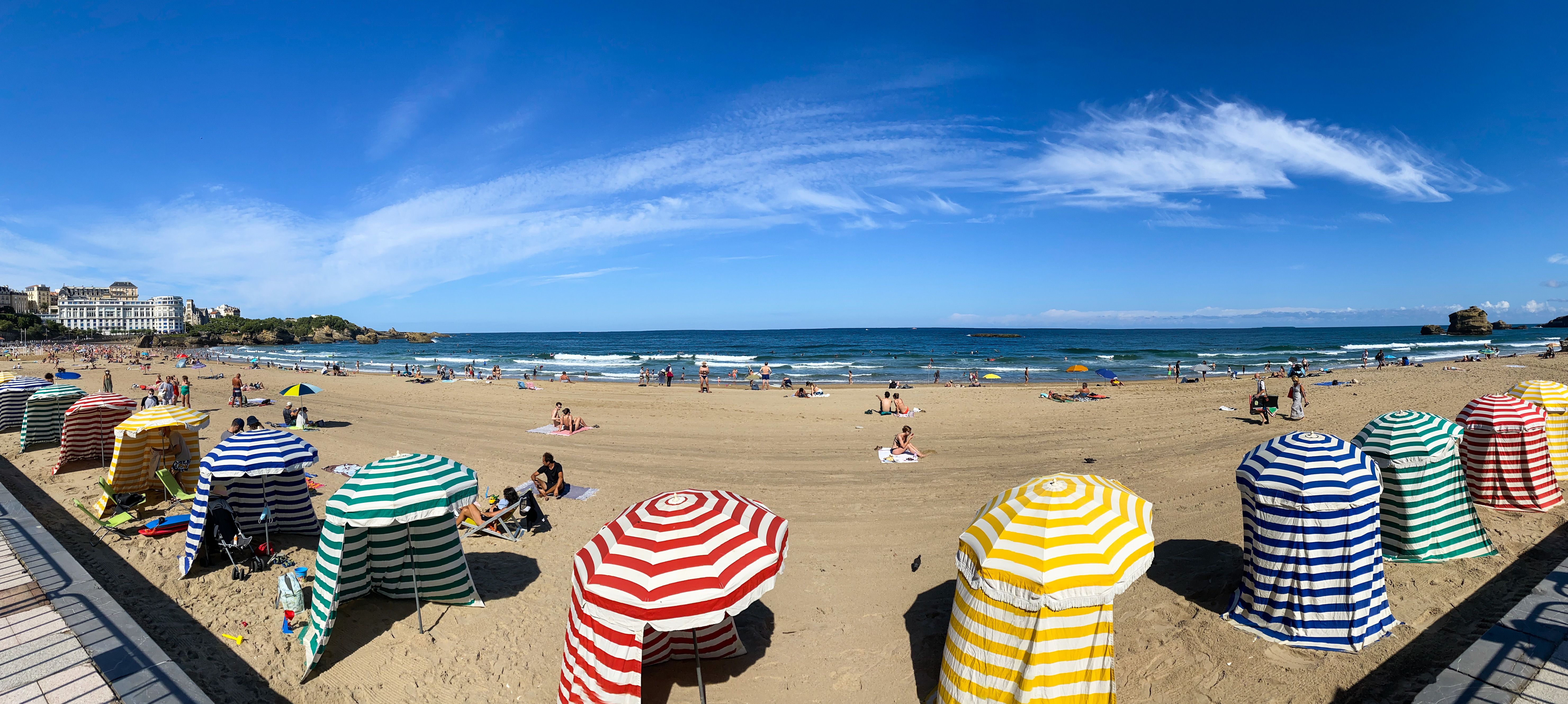 Scena di una spiaggia con rocce e mare azzurro, ombrelloni e casette sulla spiaggia, cielo limpido sopra.